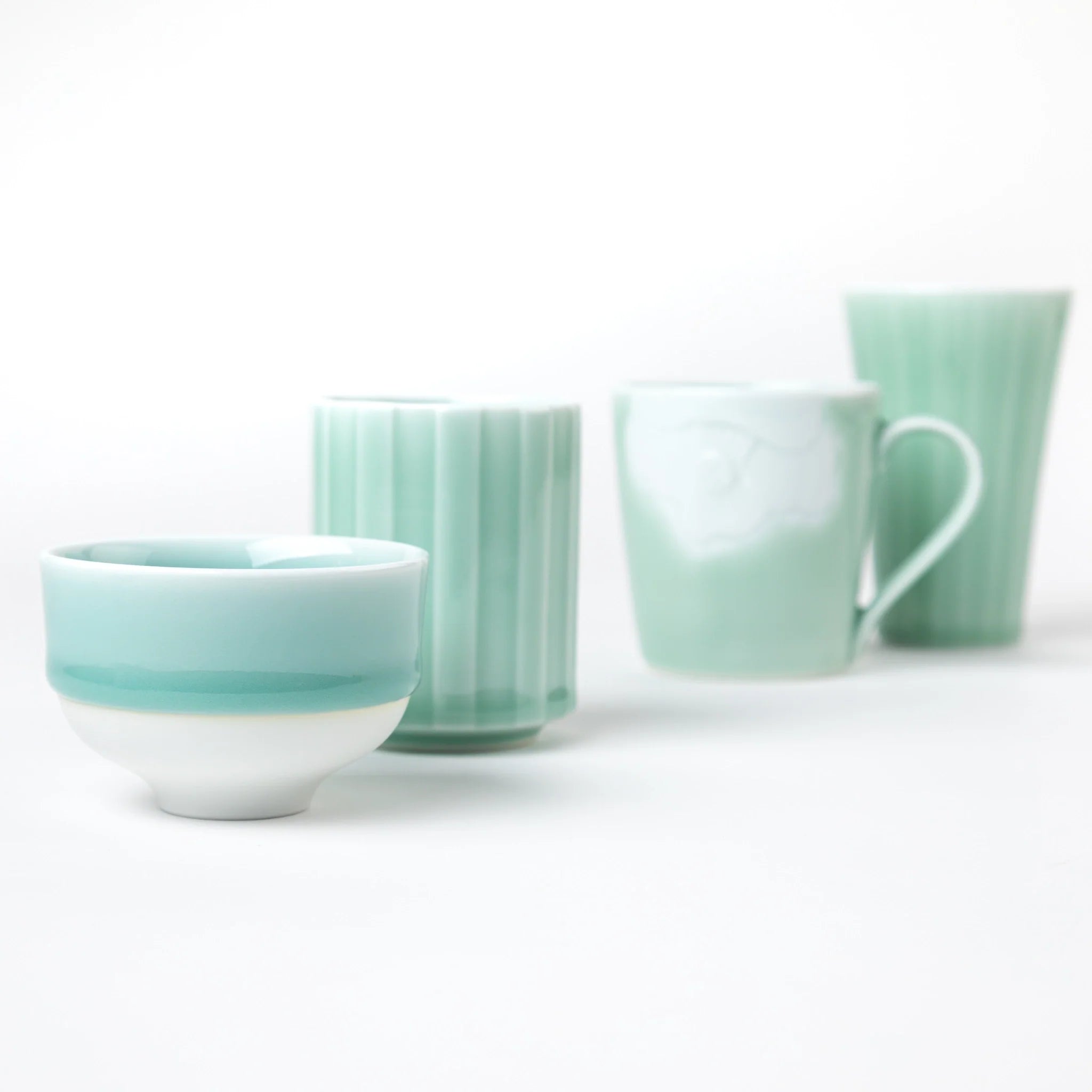 A collection of blue celadon ceramic pieces, including a tea cup, mug, and tumbler, arranged elegantly against a white background, showcasing delicate craftsmanship and a serene color palette.