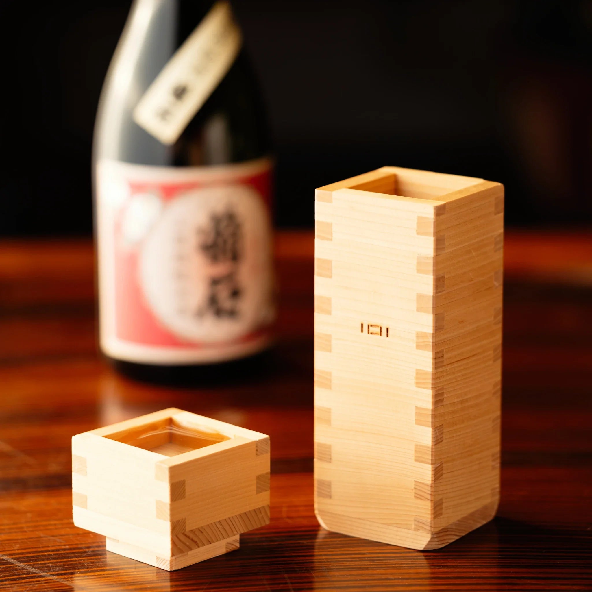 A wooden sake set featuring a small square ochoko cup and a tall tokkuri bottle on a polished wooden table with a sake bottle in the background.