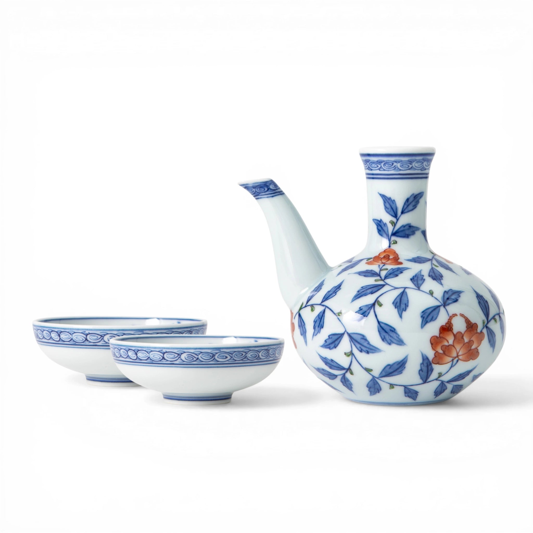 A Gen-emon Kiln Arita porcelain sake set comprising one tokkuri with a rounded body and narrow neck and two sakazuki, all decorated with a continuous cobalt blue karakusa vine motif and iron red flowers with a wave scroll border at the rims, photographed against a white background.