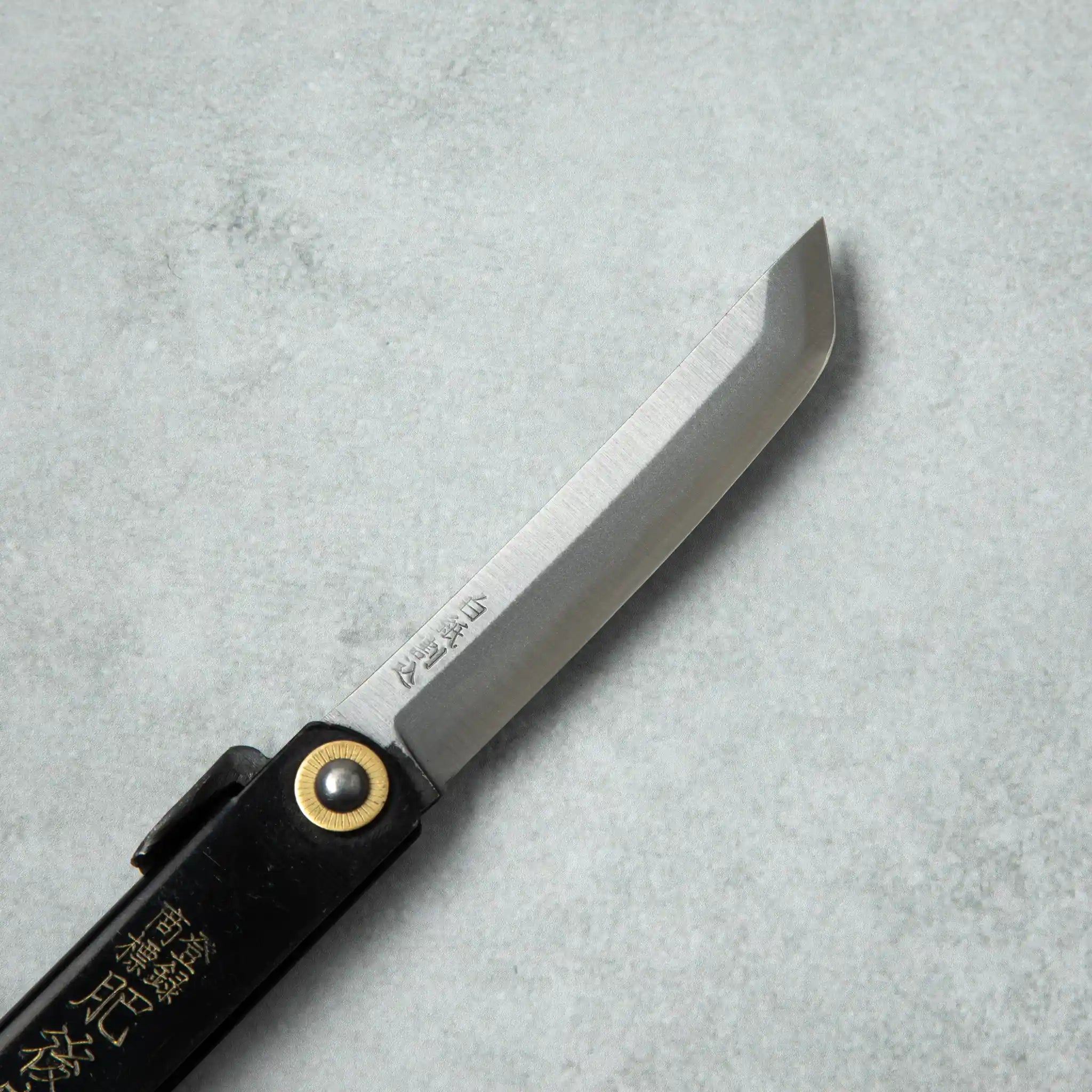 Side view of the blade of a Higonokami White Steel Sword knife, displaying its sharp edge and traditional brass pivot attached to a black handle.