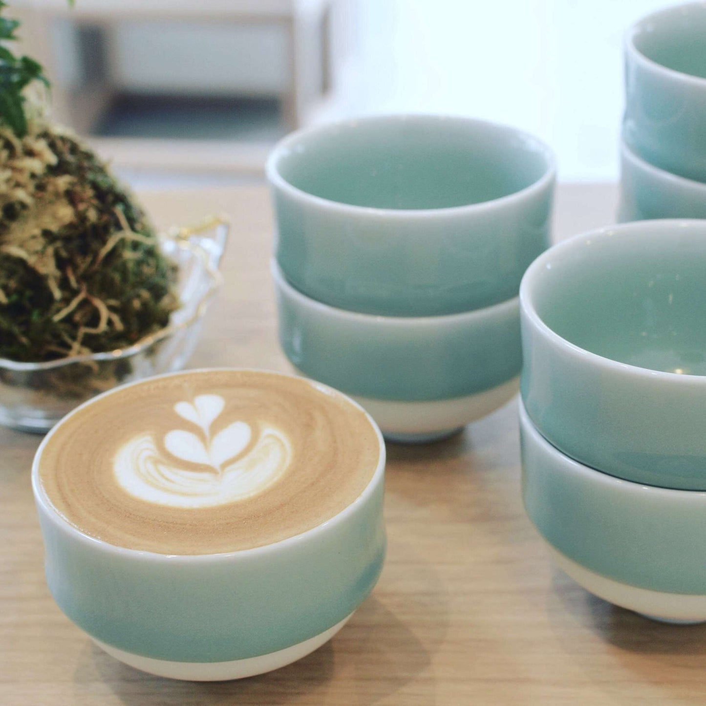 A Nabeshima celadon porcelain tea cup filled with latte art, placed next to a stack of similar cups, emphasizing its versatility for different beverages.