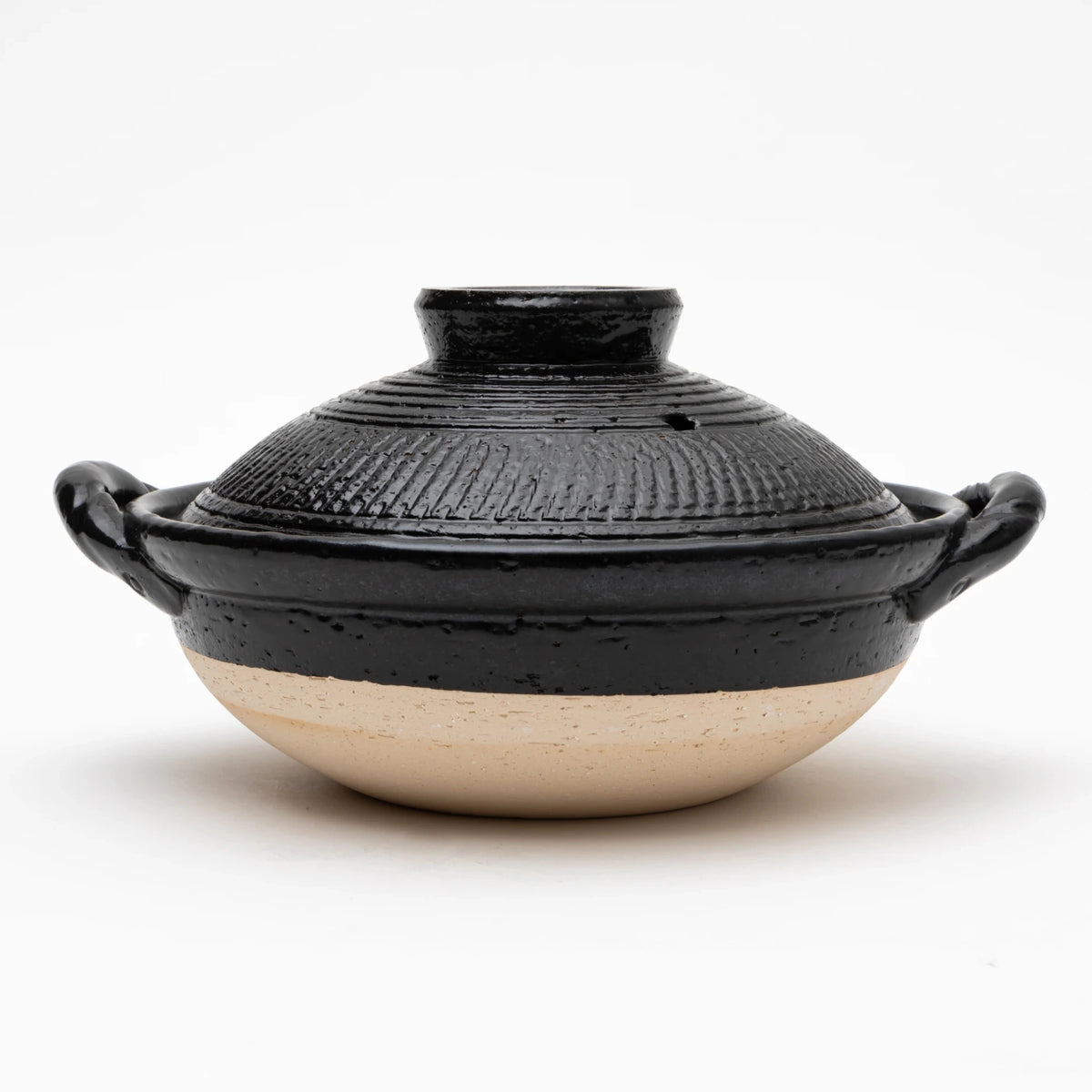 Front view of the Sajitouki Iga ware tobikanna donabe in black, showing a domed lid with fine parallel chattering tool grooves under black glaze, looped side handles, and an unglazed pale clay lower body.