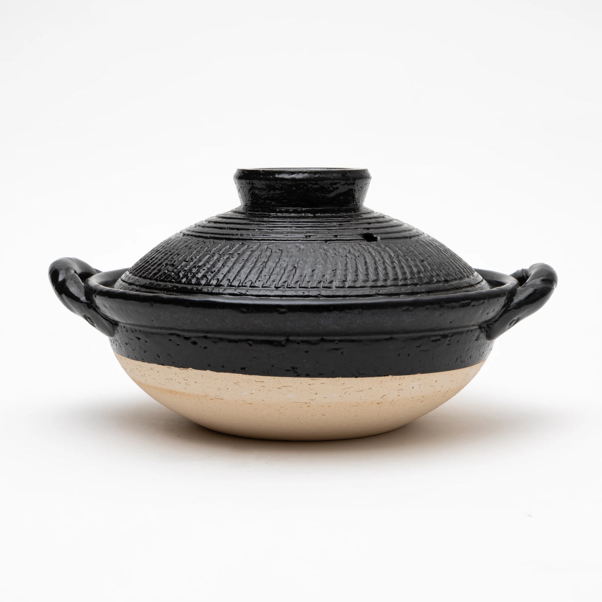 Three-quarter view of the Sajitouki Iga ware tobikanna donabe in black, showing a domed lid with fine parallel chattering tool grooves under black glaze, looped side handles, and an unglazed pale clay lower body.