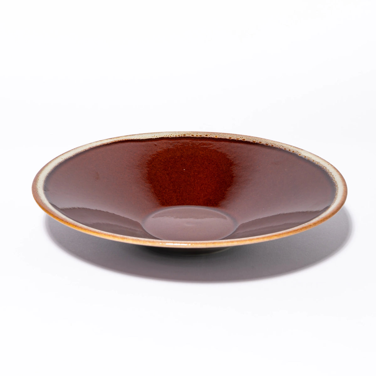 Three-quarter view of the Tsukamoto Mashiko ware SABI flat bowl large in amber glaze, showing a wide shallow form with deep iron red glaze at the center transitioning to pale grey-cream at the rim with amber iron spotting at the edge.