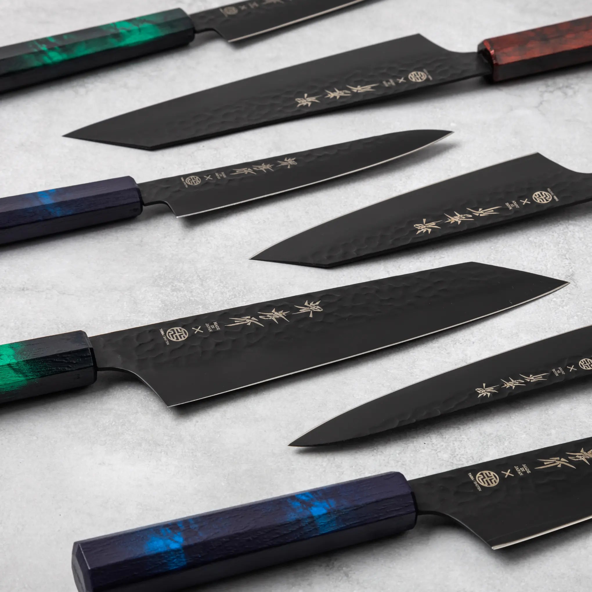 A collection of Sakai Takayuki Kurokage VG10 knives, featuring vibrant handles in green, blue, and burgundy hues, displayed diagonally with a focus on the hammered black blades and engraved kanji.
