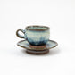 Side view of a blue and white glazed ceramic espresso cup with matching saucer, showcasing the rounded form and earthy base.