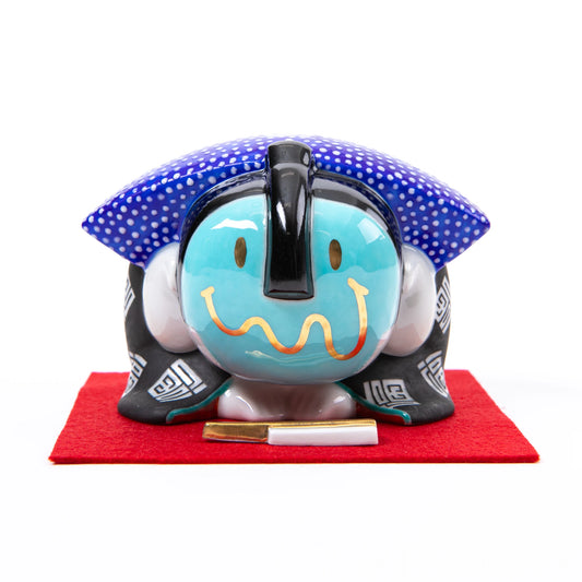 A front-facing view of the bowing Fukusuke doll, with its head tilted down, showing the glossy black hair, blue polka-dot headscarf, and a small decorative accessory on the red felt mat.