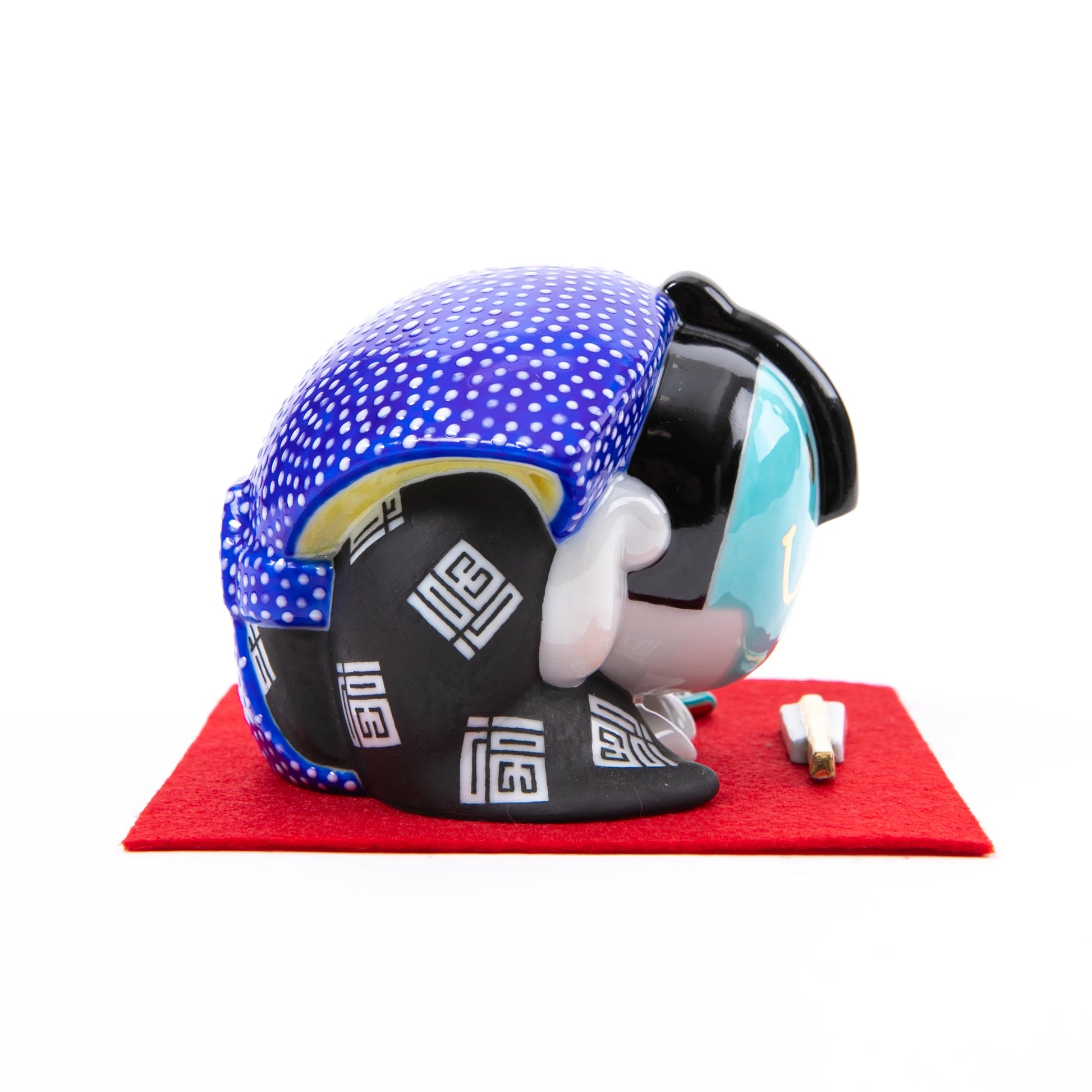 A side view of the Kutani Fukusuke doll in a deep bowing position, dressed in a black kimono with white geometric patterns and a blue polka-dot headscarf, placed on a red felt mat.