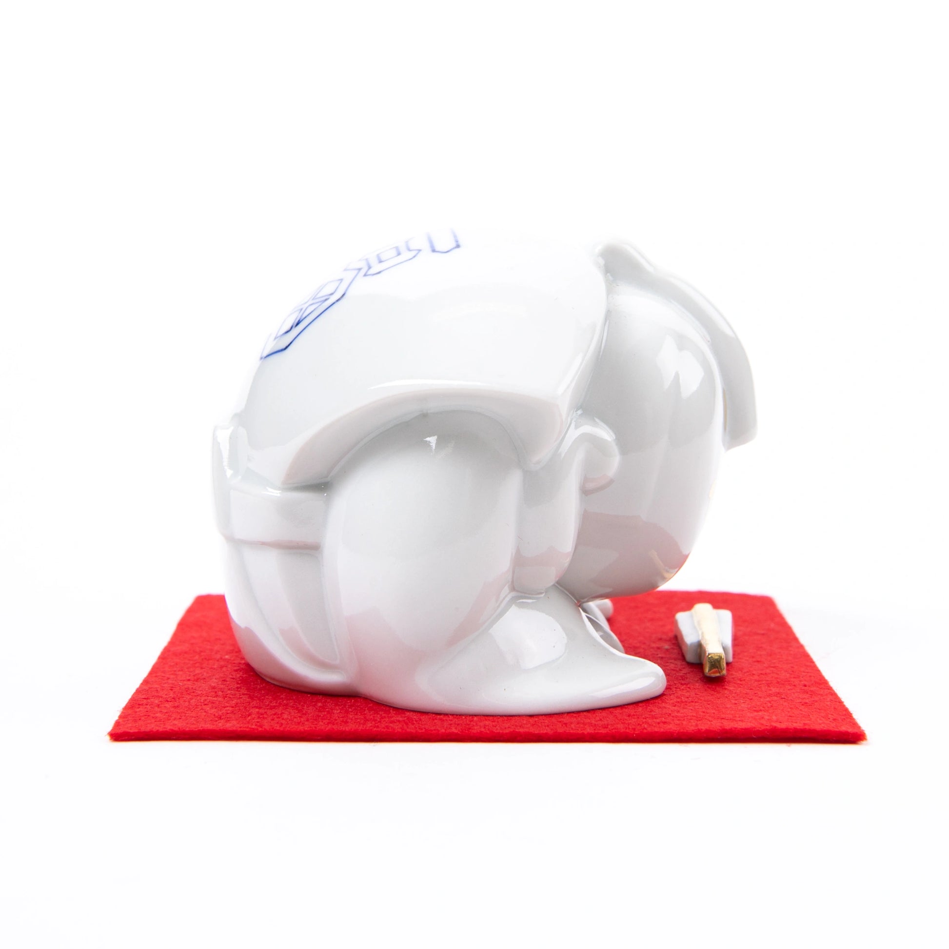 Side view of the Fukusuke Sometsuke doll, fully dressed in white with delicate gold facial features, bowing on a red mat with a decorative paper talisman.