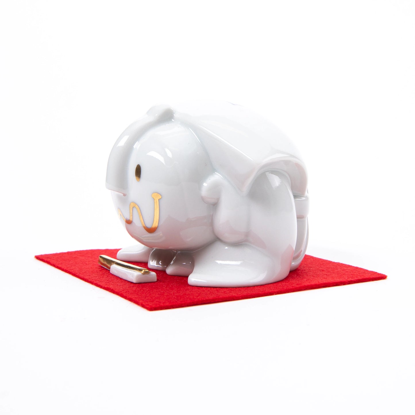 Another side view of the Fukusuke Sometsuke doll, fully dressed in white with delicate gold facial features, bowing on a red mat with a decorative paper talisman.