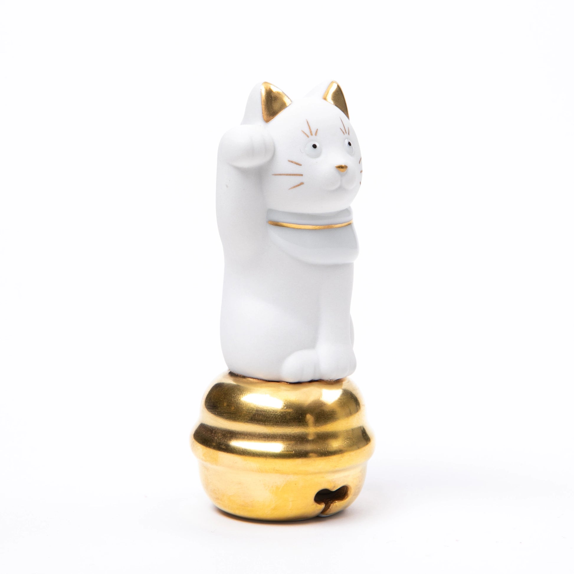 A white porcelain Maneki Neko figurine with gold accents, raising its right paw while sitting atop a polished golden bell.