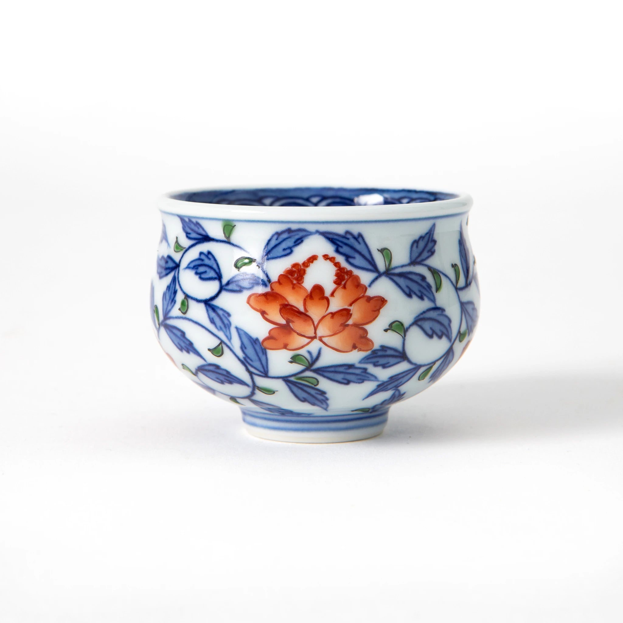 A Gen-emon Kiln Arita porcelain sake cup with a rounded body, deep cobalt blue interior, and hand painted exterior featuring a continuous cobalt blue karakusa vine motif with iron red peony flowers and green leaf accents, with a thin line border at the foot, photographed against a white background.
