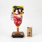 Traditional decorative hagoita paddle featuring a 3D figure of a woman in a red kimono with floral details, holding an umbrella, displayed on a wooden stand with an accompanying wooden name plaque.