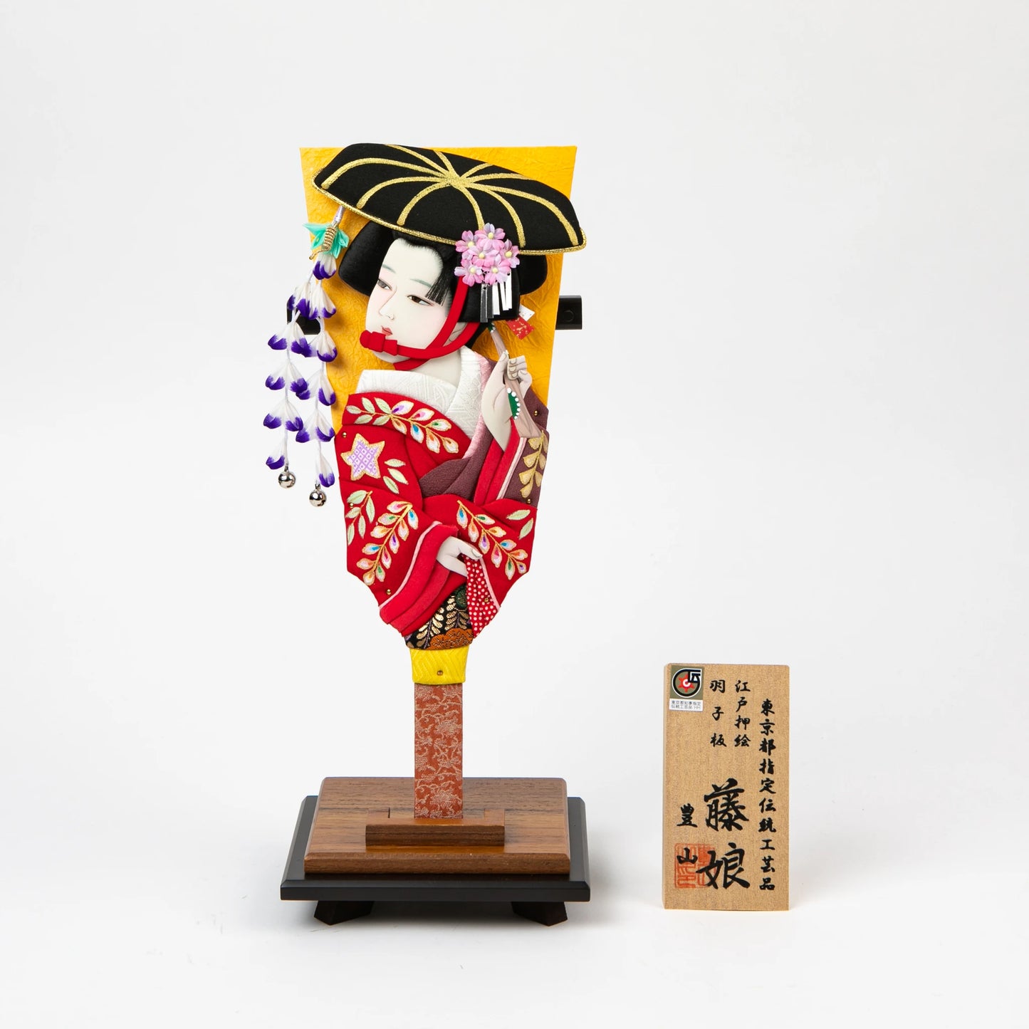 Traditional decorative hagoita paddle featuring a 3D figure of a woman in a red kimono with floral details, holding an umbrella, displayed on a wooden stand with an accompanying wooden name plaque.