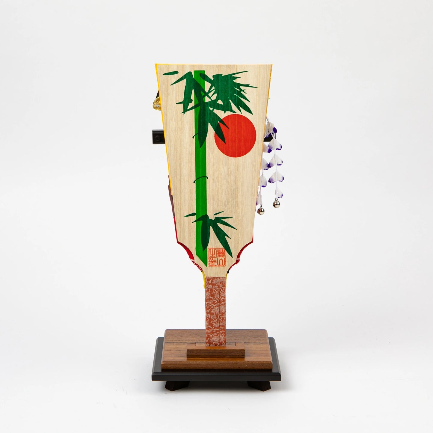 Back of a decorative hagoita paddle featuring a painted green bamboo and red sun motif on a natural wood background, displayed on a wooden stand.