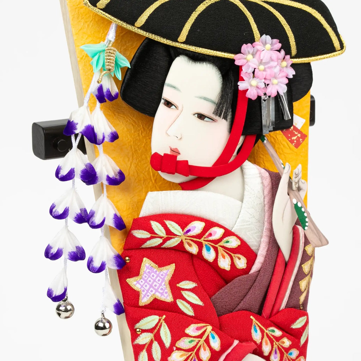 Close-up of a 3D decorative hagoita paddle showing a woman’s face in traditional makeup, wearing a red kimono and detailed hair ornaments with purple and white hanging elements.