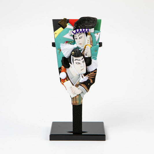 Decorative paddle featuring layered fabric reliefs of two kabuki actors in expressive poses, set against a vivid background.