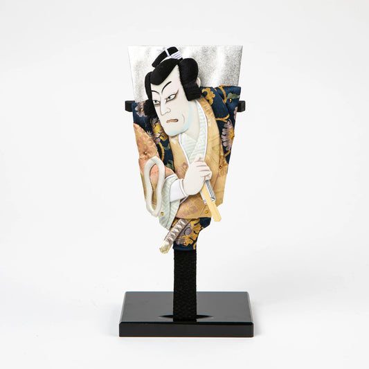 A traditional Japanese hagoita paddle featuring a male kabuki actor in a gold and navy brocade kimono, gripping a short sword.