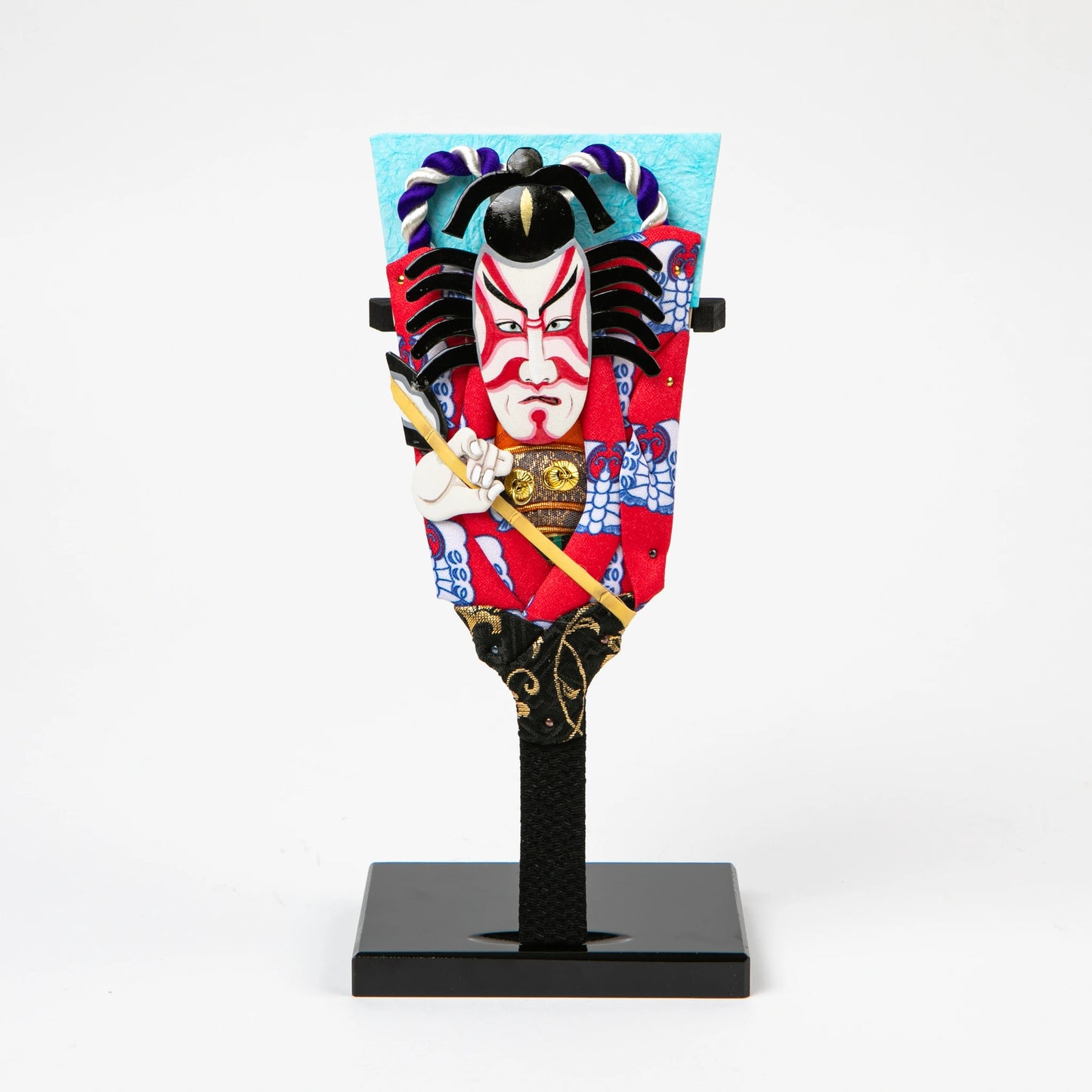 A Kabuki-inspired decorative paddle featuring a bold red costume, dramatic kumadori face paint, and intricate detailing, standing upright on a black lacquered base.