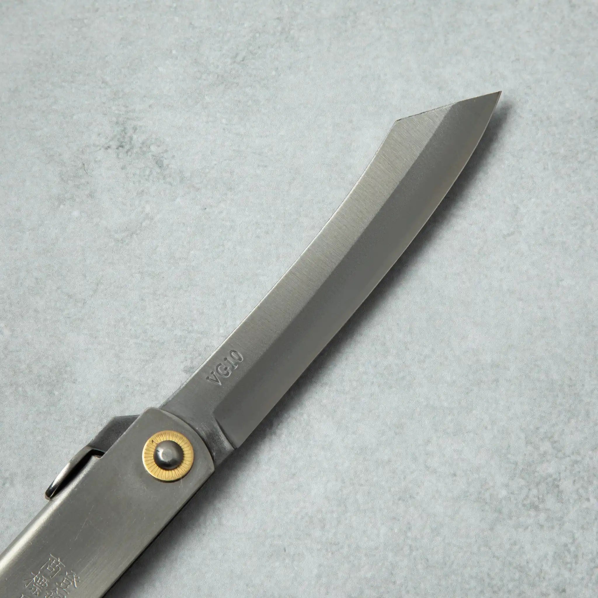 Close-up of the blade of a Higonokami VG10 stainless steel knife, featuring a curved edge and the VG10 marking near the brass pivot.