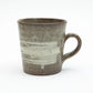 A large Shigaraki ware mug by Hinomigama with a speckled dark grey ash glaze and broad horizontal white hakeme brushstroke slip across the body, with a hand formed loop handle, set against a white background.