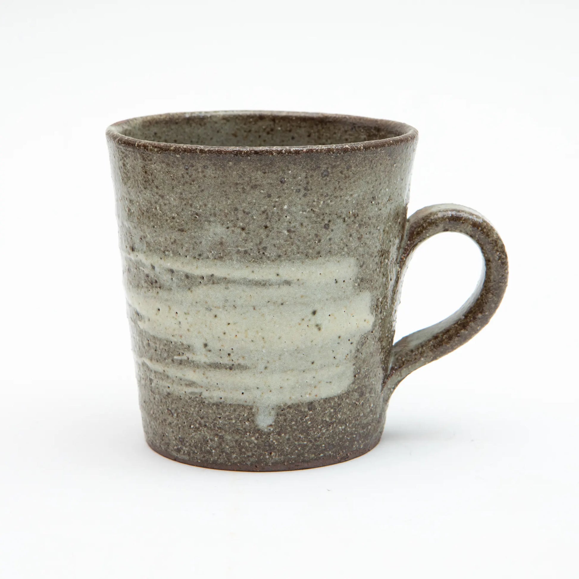 A large Shigaraki ware mug by Hinomigama with a speckled dark grey ash glaze and broad horizontal white hakeme brushstroke slip across the body, with a hand formed loop handle, set against a white background.