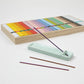 Ceramic incense holder with a lit incense stick beside the open Kosaido Hyakuraku-kou set, showing colorful sticks and packaging.