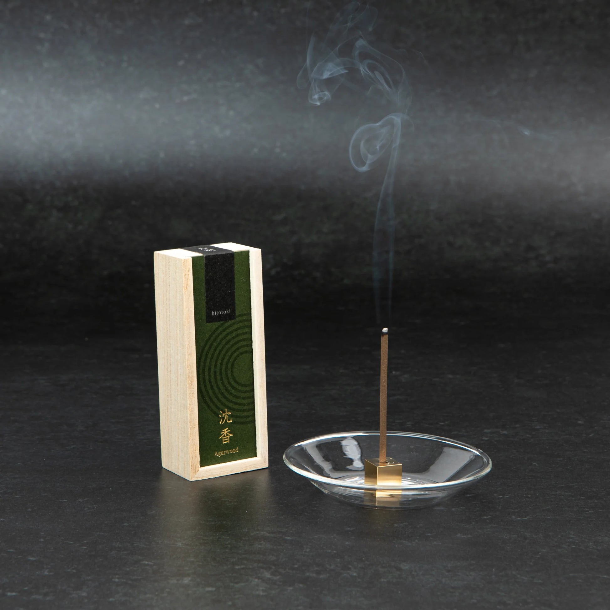 A lit agarwood incense stick gently releasing smoke from a brass holder on a glass dish, next to a green-labeled wooden box.