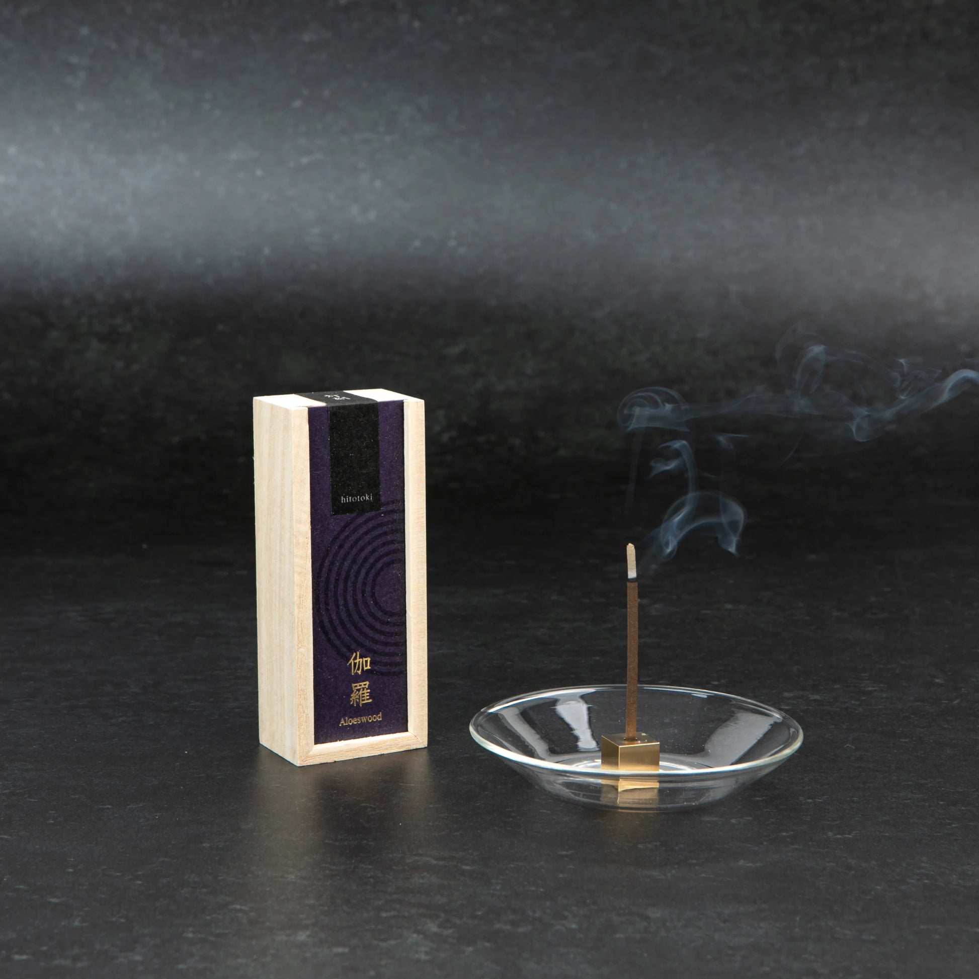 A box of Kosaido hitotoki Aloeswood incense stands upright beside a single incense stick releasing smoke in a clear glass holder, against a dark background.