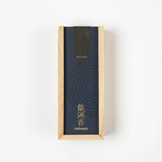 A dark blue "hitotoki" incense box labeled "Ambergris" with gold Japanese text, displayed vertically on a white background.