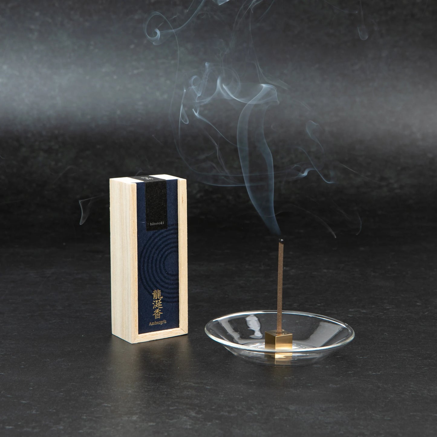 A burning incense stick releasing visible smoke beside the "Ambergris" hitotoki incense box, with a modern glass holder on a dark background.