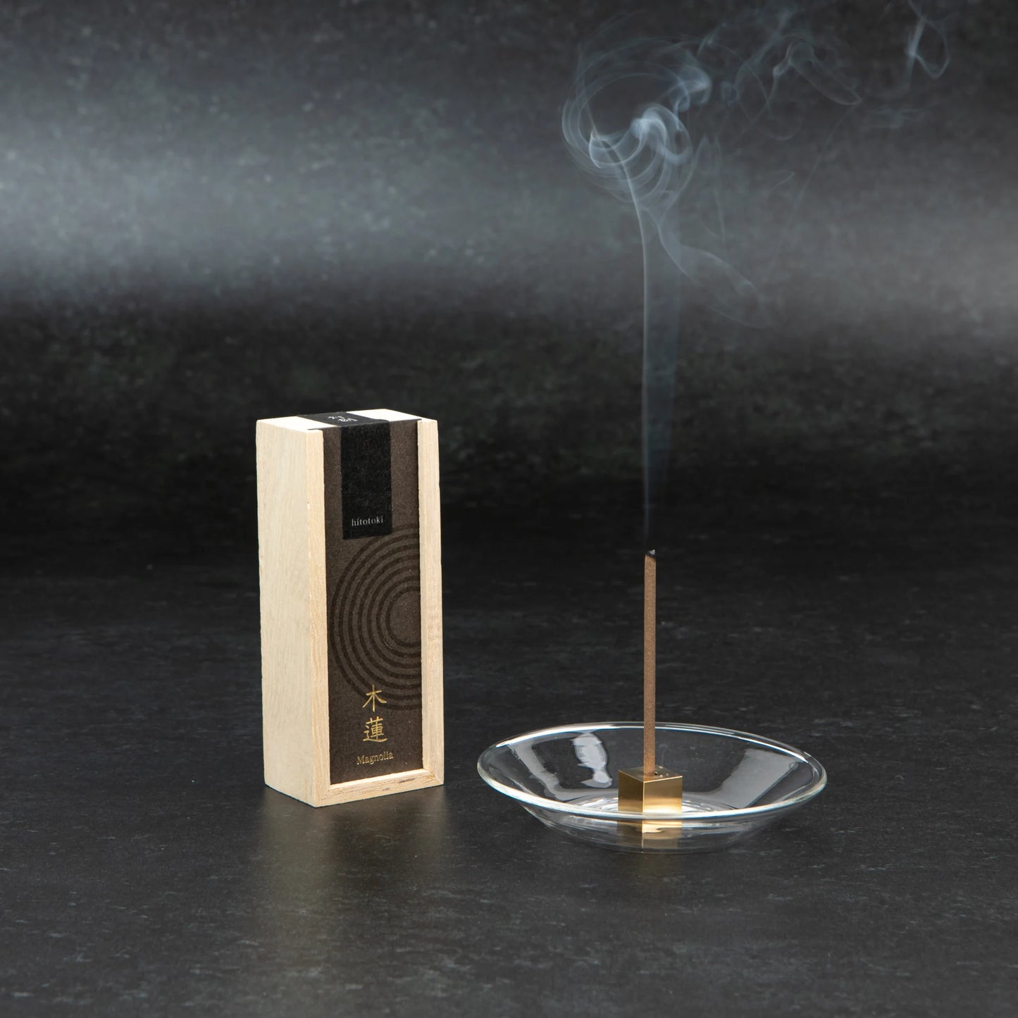 A box of hitotoki Magnolia incense standing beside a glass dish with a lit incense stick releasing a soft trail of smoke.