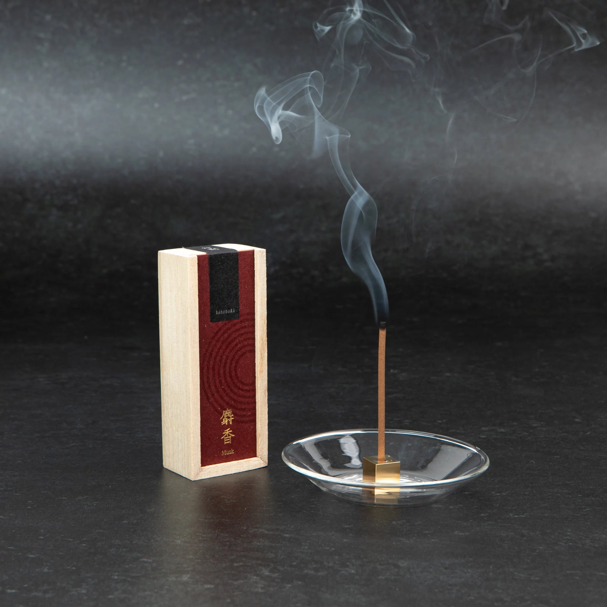 Kosaido hitotoki musk incense burning in a glass holder with visible smoke, next to the upright wooden box, set against a dark backdrop.