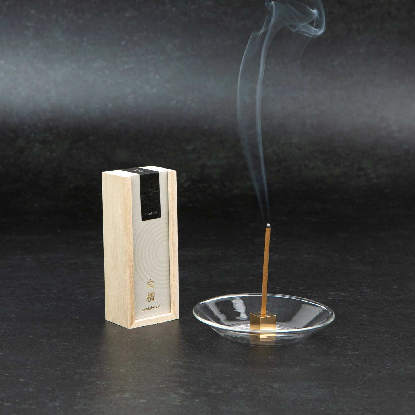Sandalwood incense stick burning in a glass holder beside a light wooden box with black and beige label on a dark stone background