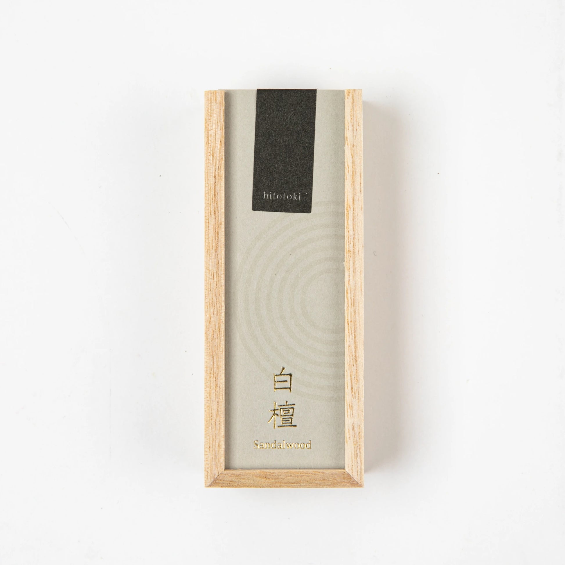 Sandalwood incense stick burning in a glass holder beside a light wooden box with black and beige label on a dark stone background