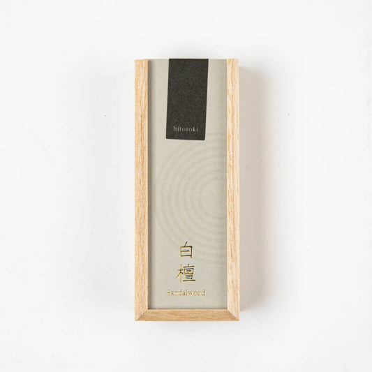 Sandalwood incense stick burning in a glass holder beside a light wooden box with black and beige label on a dark stone background