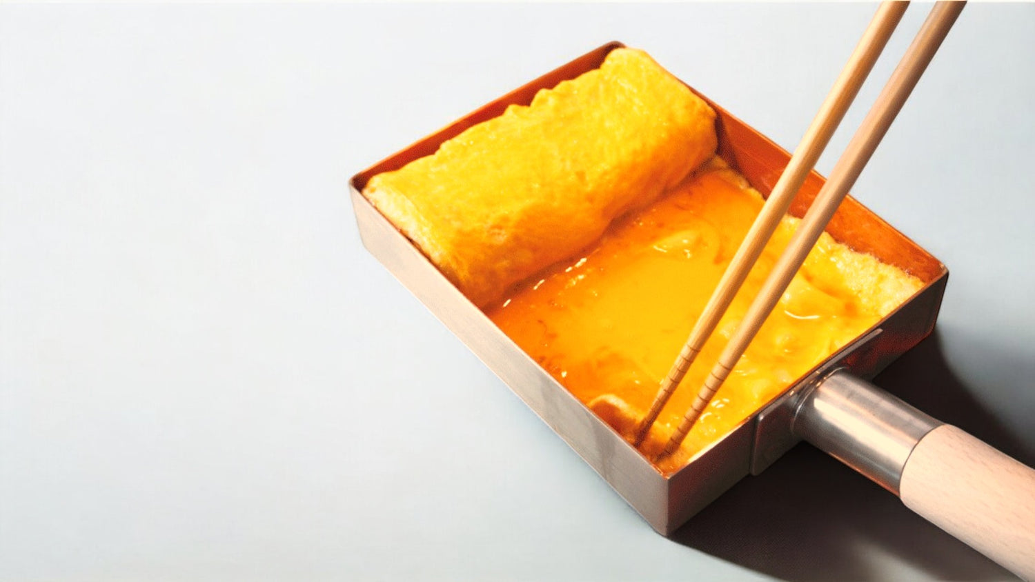 Rolled omelet Tamagoyaki being prepared in a rectangular copper tamagoyaki pan using chopsticks, with a wooden handle visible.