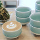 A Nabeshima celadon porcelain tea cup filled with latte art, placed next to a stack of similar cups, emphasizing its versatility for different beverages.