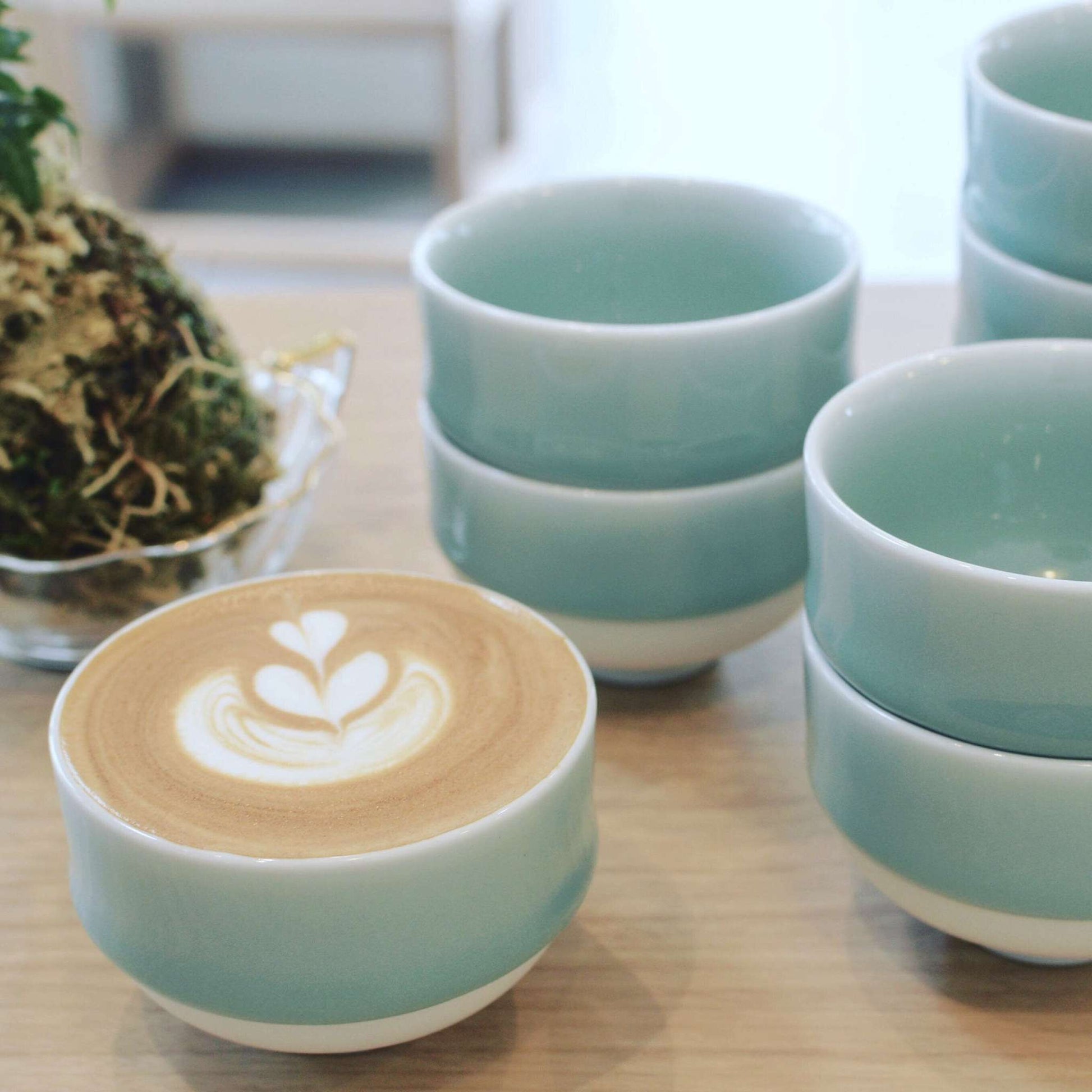 A Nabeshima celadon porcelain tea cup filled with latte art, placed next to a stack of similar cups, emphasizing its versatility for different beverages.