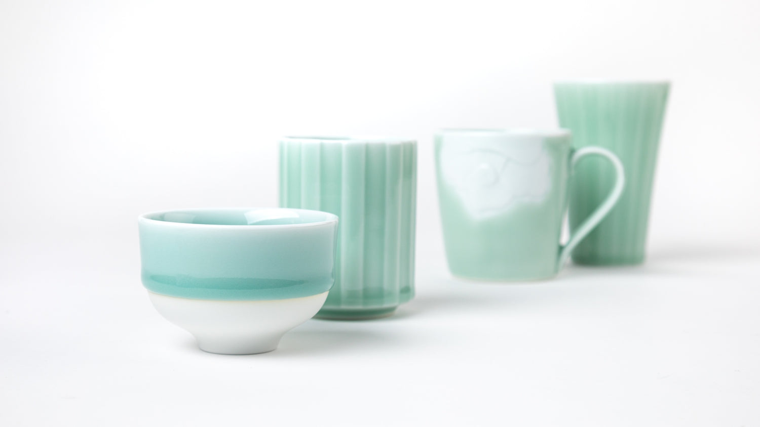 A collection of blue celadon ceramic pieces, including a tea cup, mug, and tumbler, arranged elegantly against a white background, showcasing delicate craftsmanship and a serene color palette.