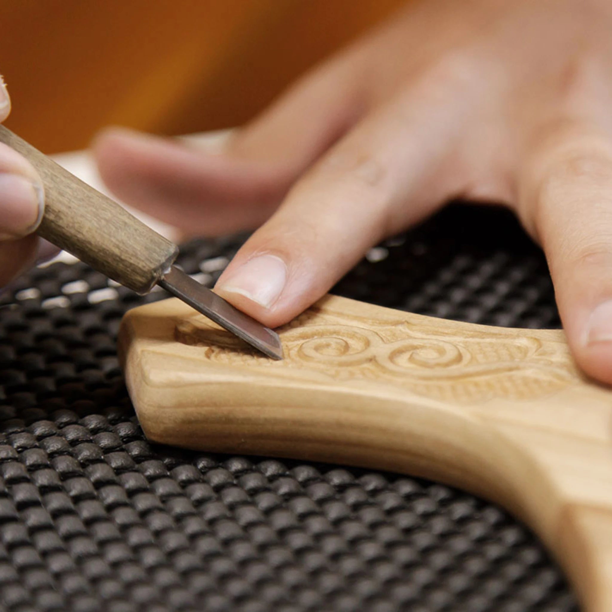 Hand using a small wood carving knife on a wooden object with intricate carvings.