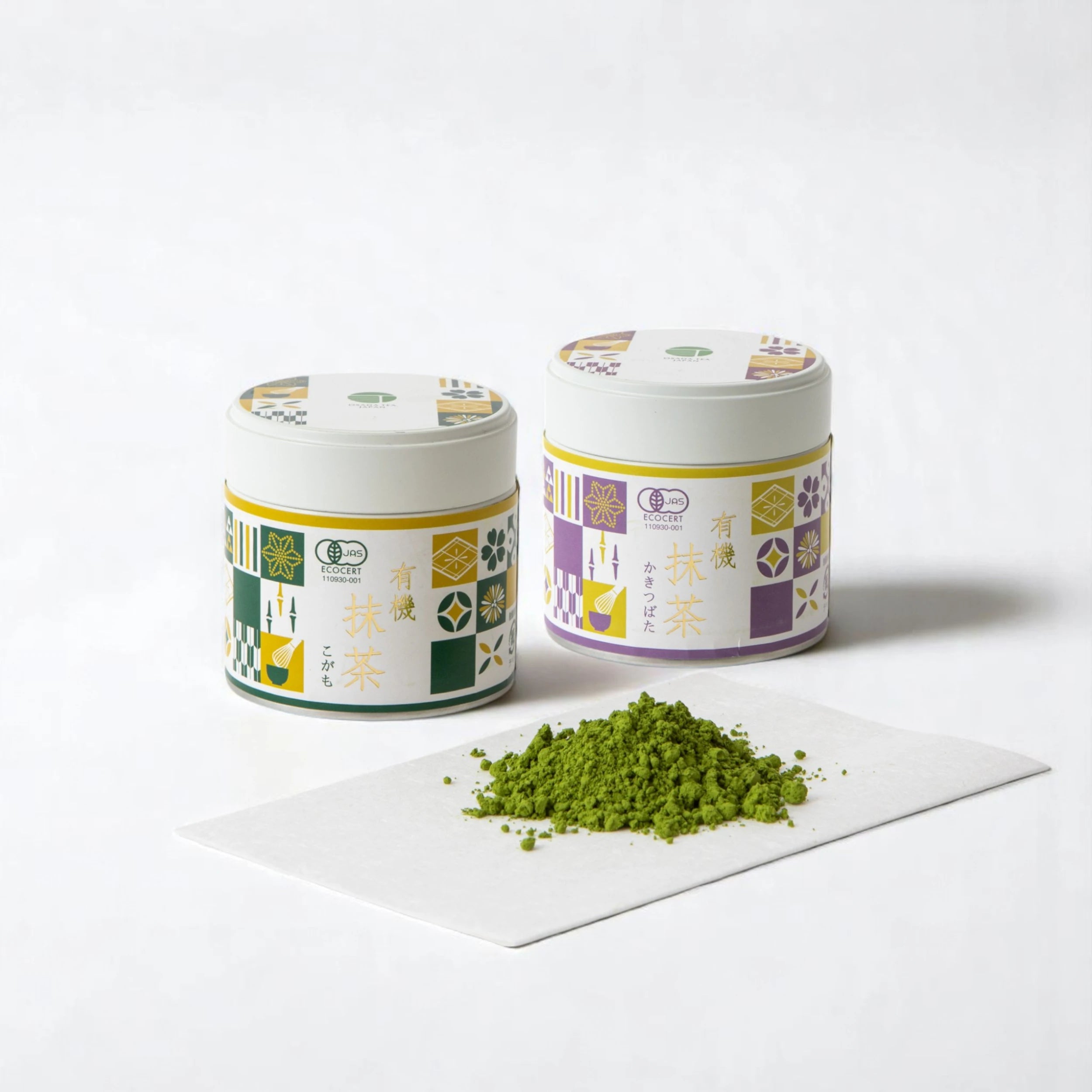 Two Osada Tea organic matcha tins, OG Matcha Kakitsubata in a purple patterned tin and OG Matcha Kogamo in a green patterned tin, photographed side by side with a small pile of vivid green matcha powder on a white sheet, against a white background.