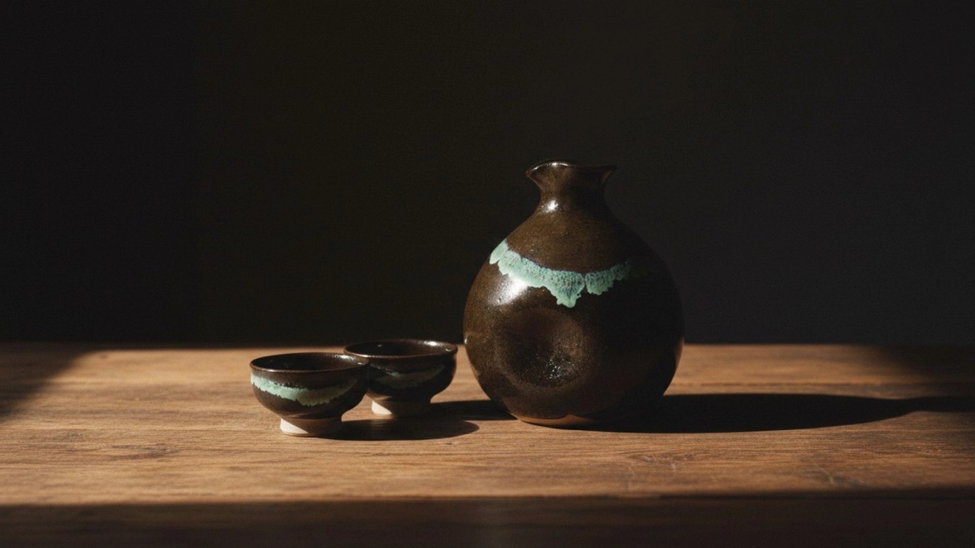 Ryumonji ware black ceramic sake carafe and cups on a wooden surface with a dark background