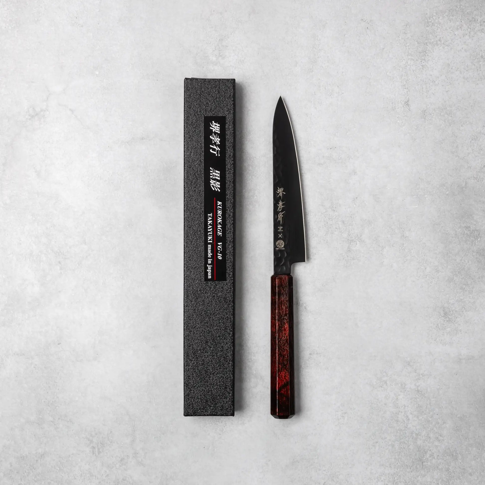 A Sakai Takayuki Kurokage VG10 petty knife with a hammered black blade and a luxurious burgundy resin octagonal handle, displayed alongside its black textured packaging on a neutral background.
