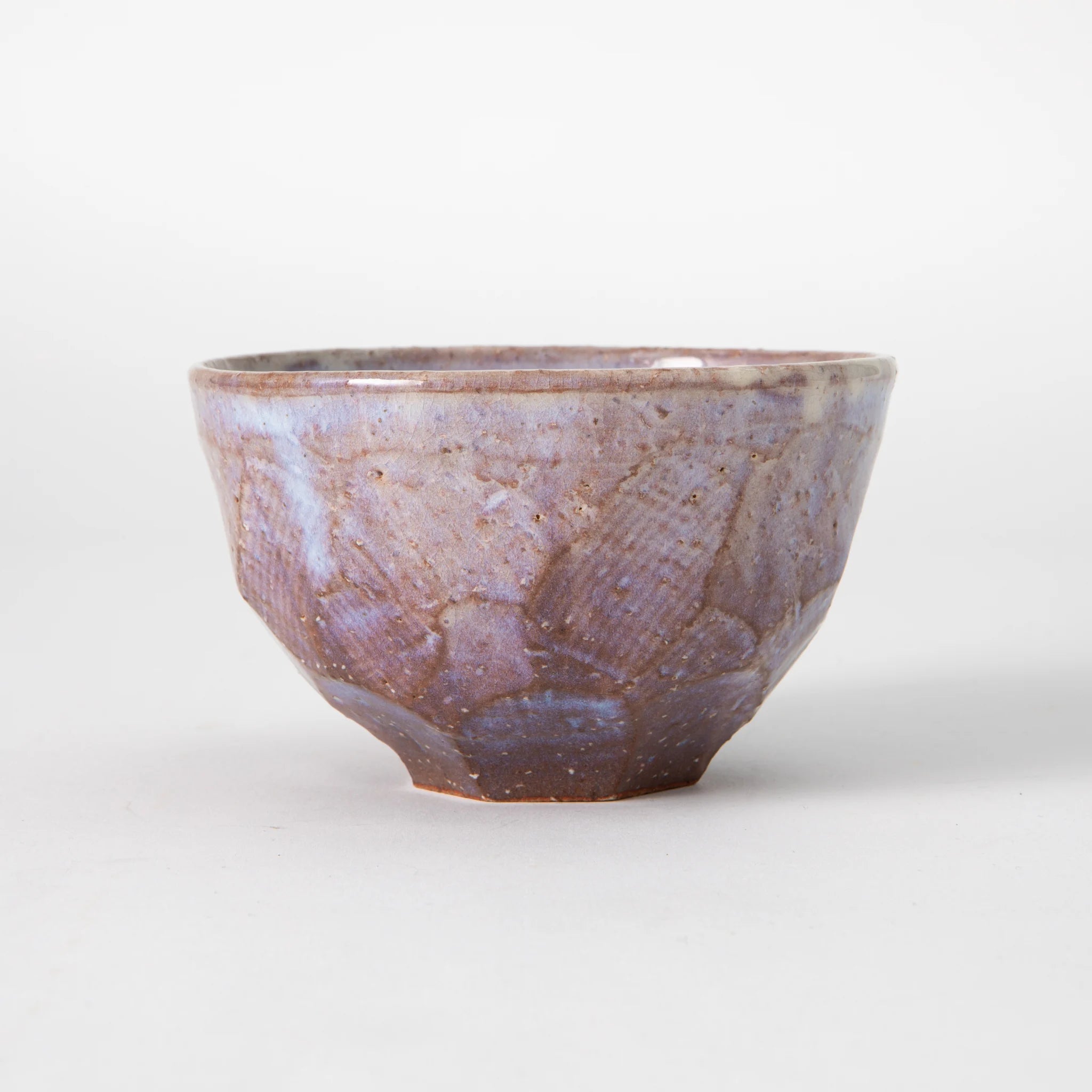 A Hagi ware matcha bowl by Senryuzan with broad Misogi carved exterior facets beneath a white slip and translucent glaze in dusty lavender and muted rose tones, with warm iron clay visible through the glaze at the ridges, photographed against a white background.