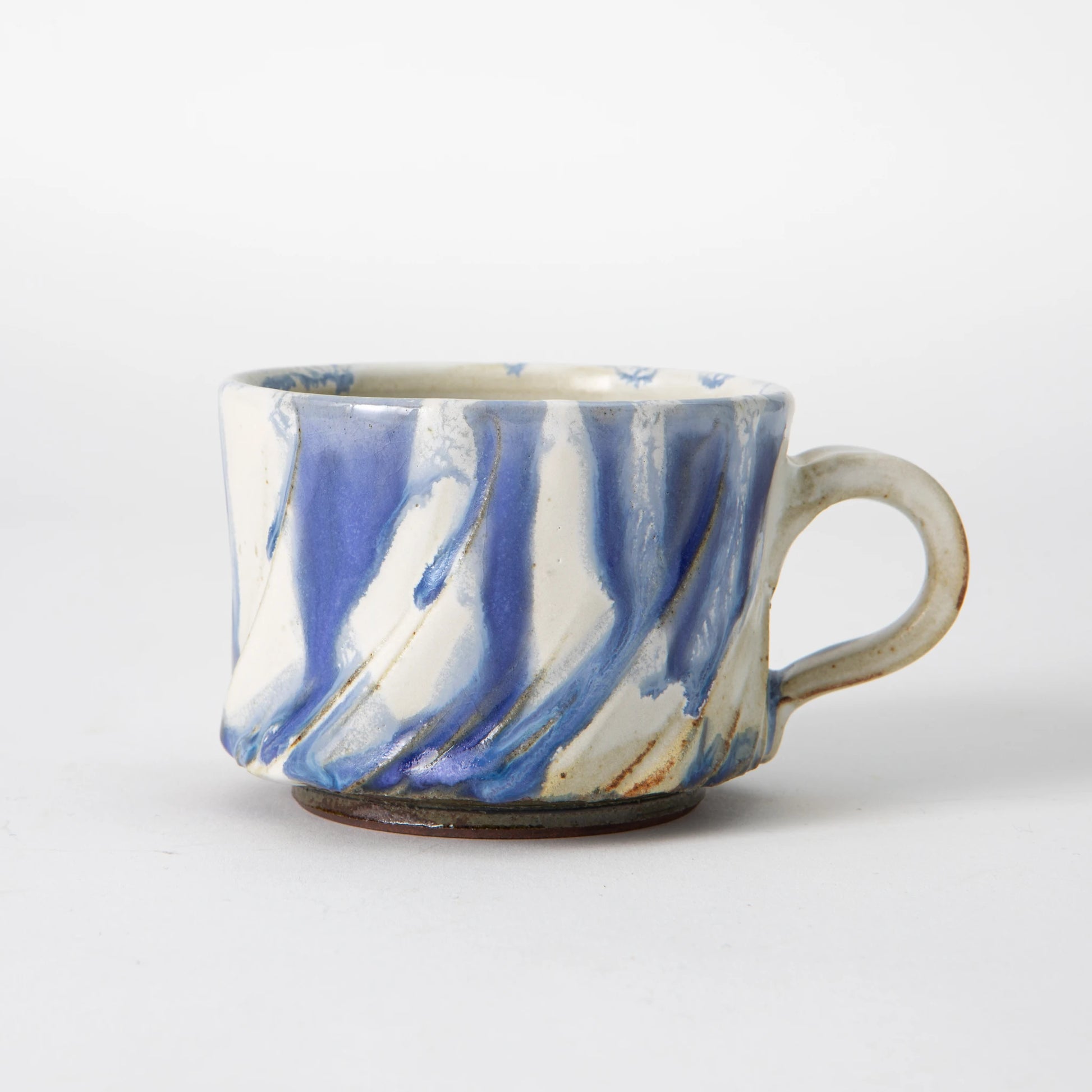A wide, low Tamba ware mug by Shozo Gama with diagonal hand carved facets covered in soft periwinkle blue and chalk white ash glaze with fine iron lines along the ridges, a pale loop handle, and an unglazed dark foot, photographed against a white background.