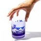 A hand reaching out to hold the blue whiskey glass adorned with Mount Fuji and sakura designs, giving a sense of scale