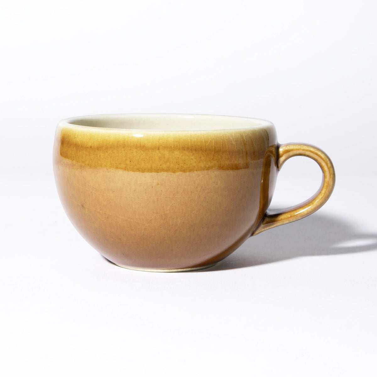 Side view of the Tsukamoto Mashiko ware soup cup in amber glaze, showing a wide rounded form with a loop handle, glaze transitioning from pale cream at the rim through golden amber to rosy brown at the base, with fine crazing across the surface.