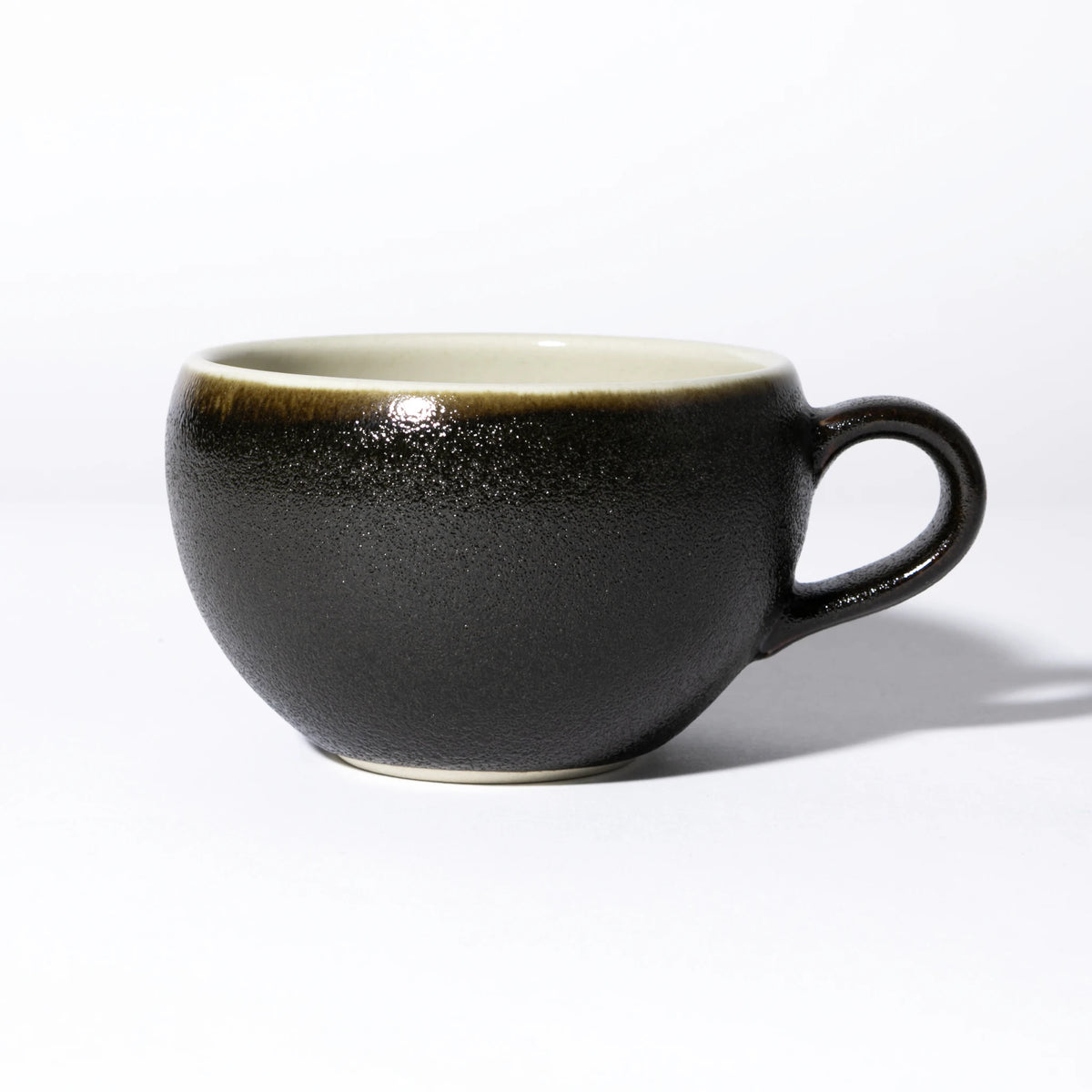 Side view of the Tsukamoto Mashiko ware soup cup in black glaze, showing a wide rounded form with a loop handle, heavily textured granular black glaze across the body transitioning to warm amber at the rim, and a pale cream interior.
